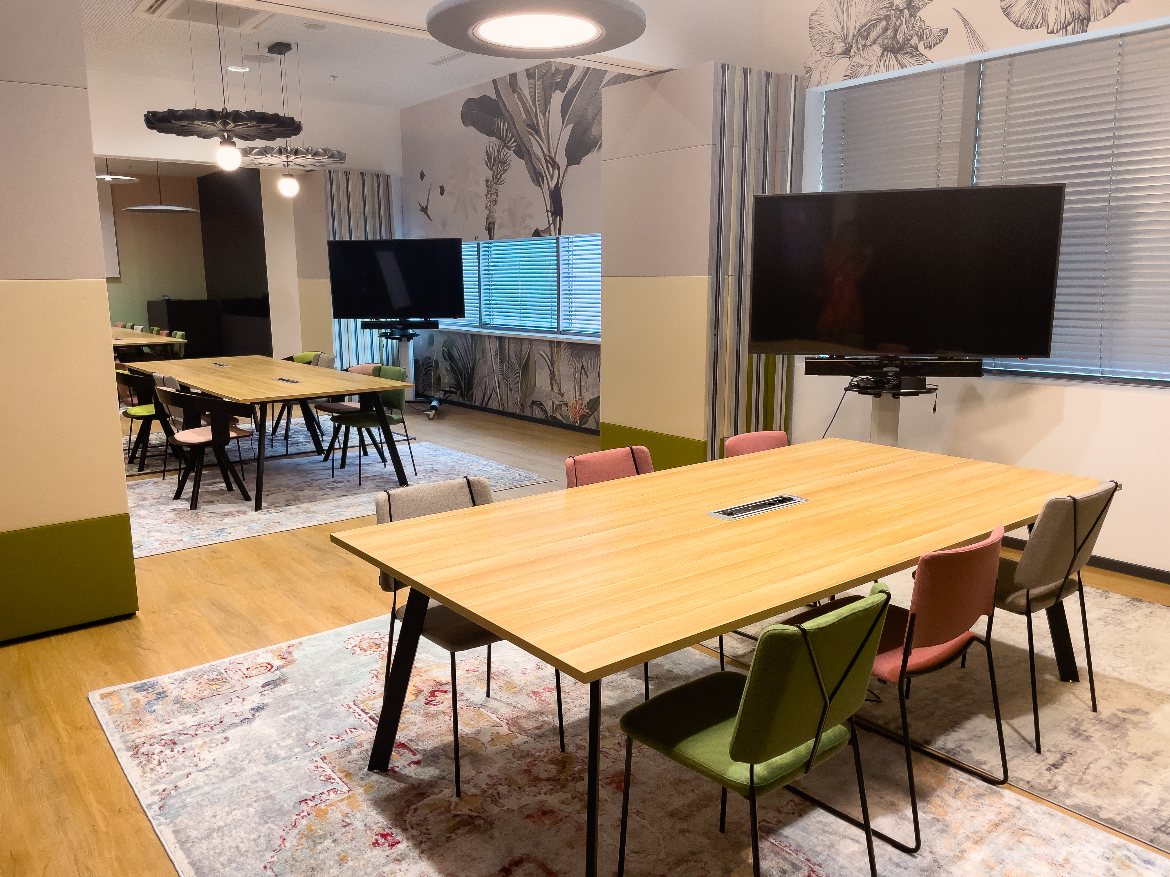 Videokonferencia-ozvucenie-priestorov Meeting room with a wooden table, colourful chairs and a wall display with a video conferencing bar